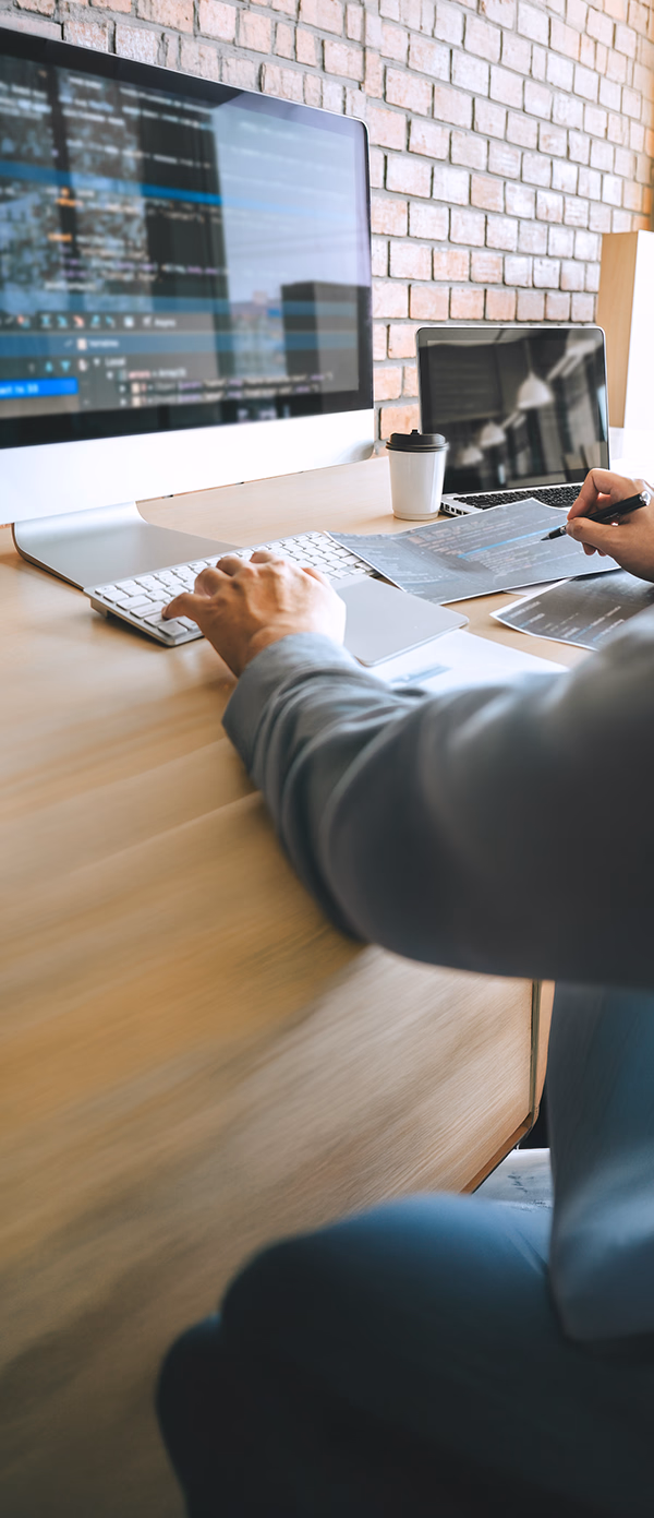 Website development service hero banner featuring a programmer coding on a large desktop monitor and reviewing project schematics at a modern desk.