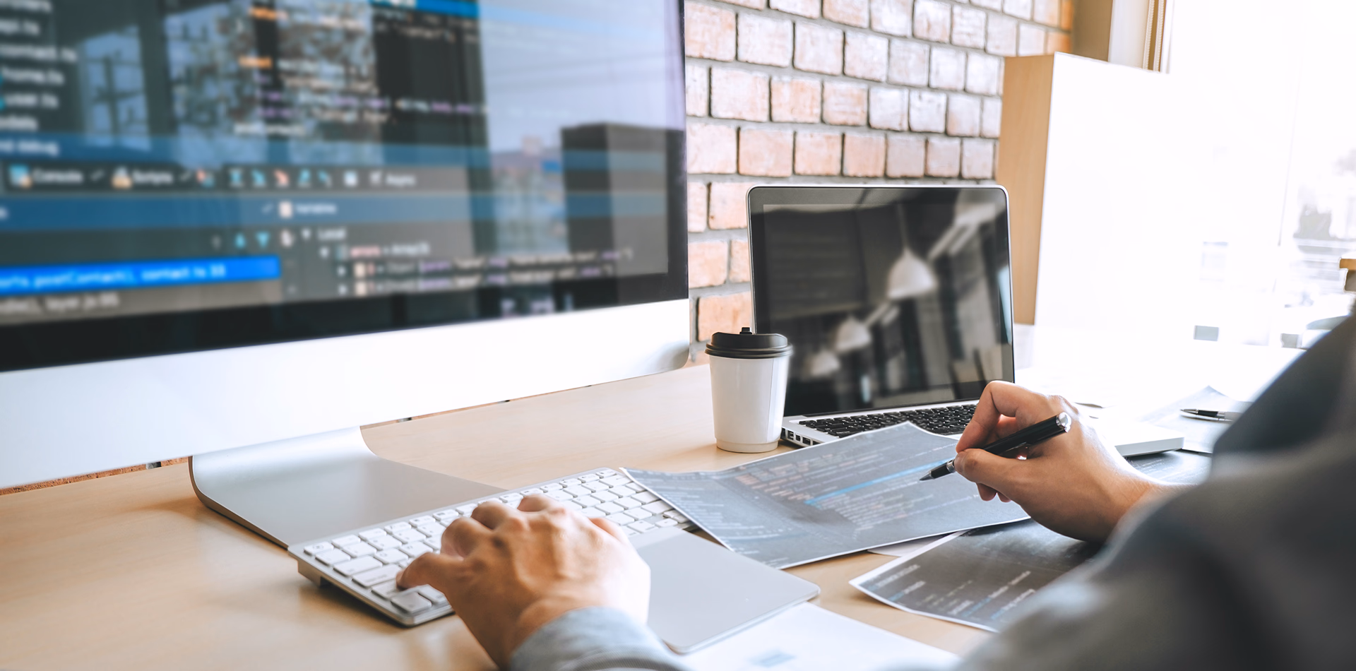 Website development service hero banner featuring a programmer coding on a large desktop monitor and reviewing project schematics at a modern desk.